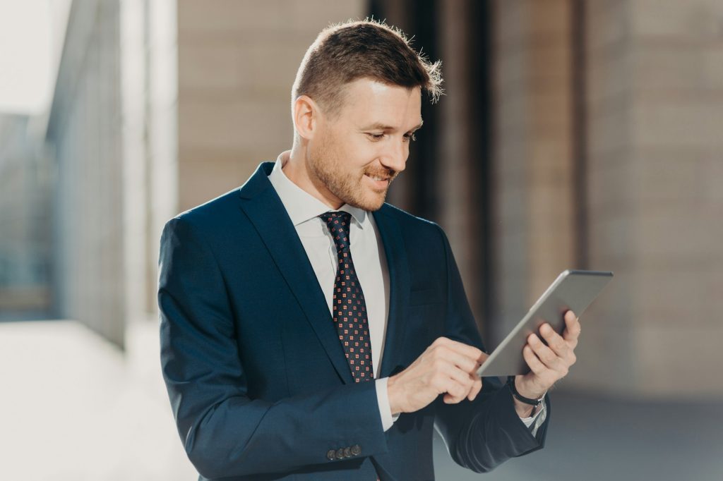 Successful male office worker dressed in elegant suit, holds modern digital tablet, updates website